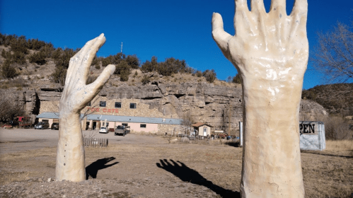 Two large, sculpted hands reaching up from the ground, set against a rocky landscape and a building in the background.