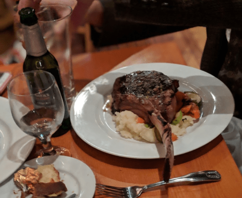 A plate with a large pork chop on mashed potatoes and vegetables, accompanied by a glass of water and a bottle.