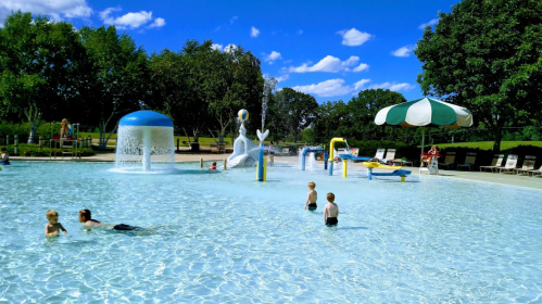 Children playing in a splash pad area with water features, surrounded by trees and blue skies.