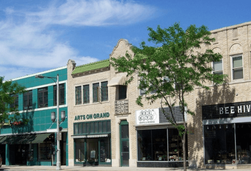 A sunny street scene featuring colorful storefronts, including "Arts on Grand" and "Bee Hive," with trees lining the sidewalk.