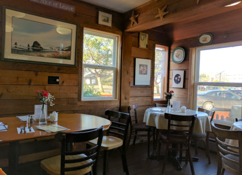 Cozy restaurant interior with wooden walls, tables set for dining, and framed artwork near large windows.