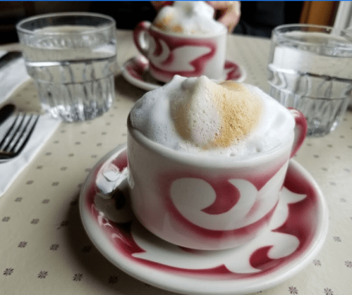 Two cups of frothy coffee on decorative saucers, with glasses of water in the background on a patterned tablecloth.