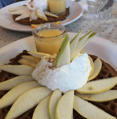 A waffle topped with sliced green apples, whipped cream, and a small cup of yellow sauce, served on a white plate.