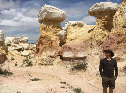 A person stands in a colorful, rocky landscape with unique rock formations under a partly cloudy sky.