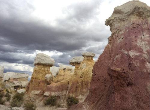 Colorful rock formations under a cloudy sky, showcasing layers of red, yellow, and white in a desert landscape.