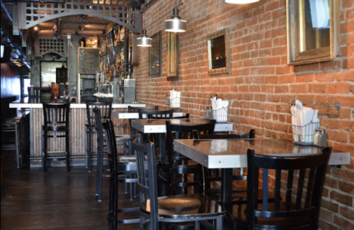 Cozy restaurant interior with brick walls, black tables, and chairs, featuring a bar in the background.