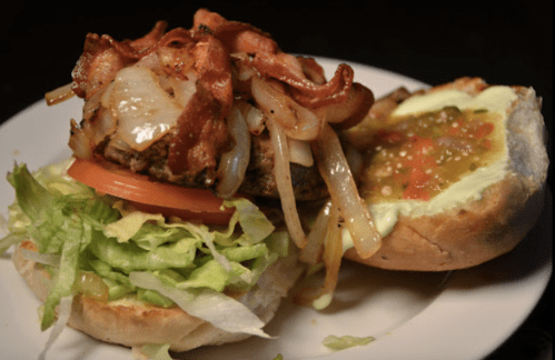 A close-up of a burger with bacon, grilled onions, lettuce, and tomato on a bun, served on a plate.