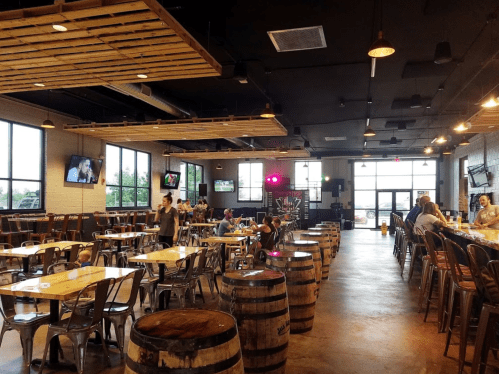A spacious bar and dining area with wooden tables, barrels, and large windows, featuring a lively atmosphere.