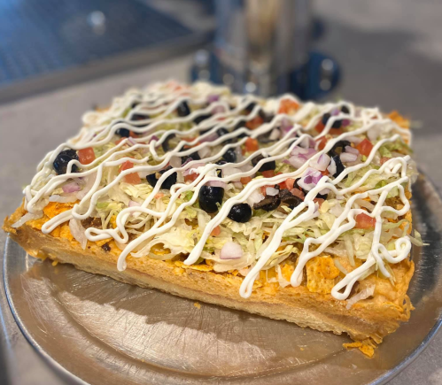 A square pizza topped with lettuce, tomatoes, olives, onions, and drizzled with white sauce on a metal tray.
