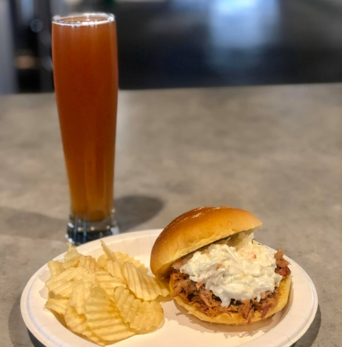 A plate with a pulled pork sandwich topped with coleslaw, served with potato chips and a tall glass of iced tea.