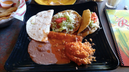 A black plate with two tacos, rice, refried beans, and an enchilada, garnished with lettuce and tomatoes.