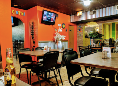 Interior of a colorful restaurant with orange walls, tables, chairs, and a TV showing a sports game.