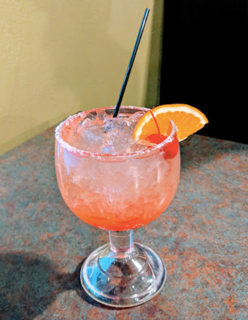 A colorful cocktail in a glass, garnished with a cherry and an orange slice, served on a textured surface.