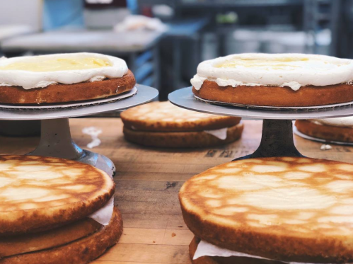 Two layered cakes on stands, one with frosting and lemon topping, in a bakery setting with blurred background.