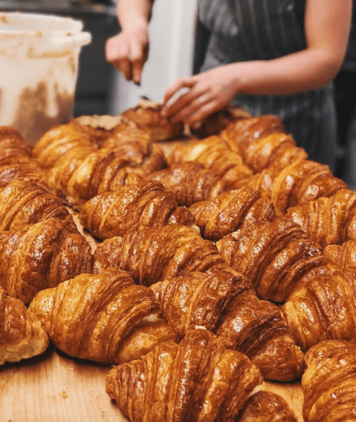 A close-up of freshly baked croissants on a wooden table, with a person in the background preparing more pastries.
