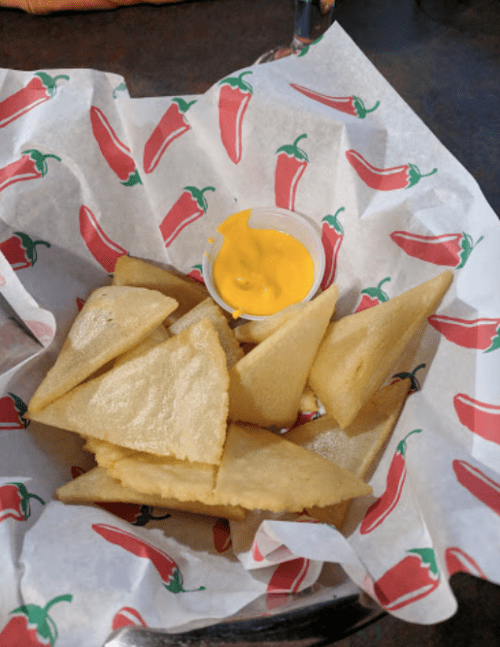 A basket of triangular corn tortilla chips with a small cup of yellow cheese dip, lined with chili pepper-patterned paper.