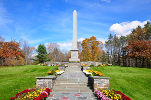 A tall white monument surrounded by colorful flowers and green grass, set against a blue sky with scattered clouds.