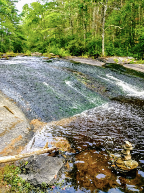 A serene river flows over rocks, surrounded by lush greenery, with a small stack of stones balanced nearby.