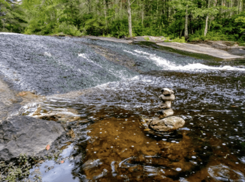 A serene river scene with a small waterfall and a stacked stone sculpture in the foreground. Lush greenery surrounds the area.