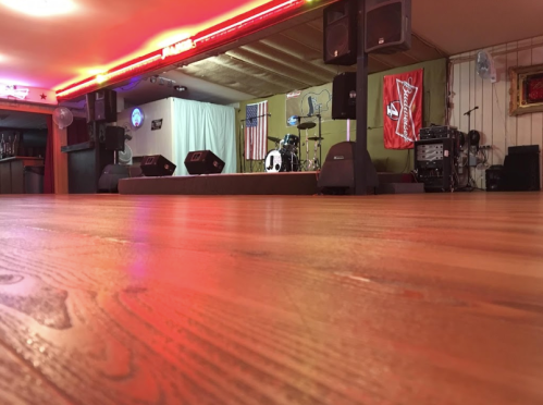 A dimly lit stage with musical equipment, flags, and red lighting, viewed from the floor level.