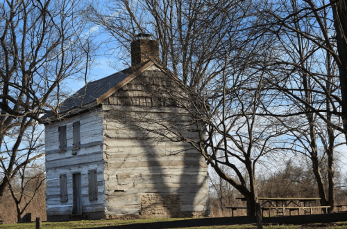 A weathered log cabin surrounded by bare trees under a clear blue sky.