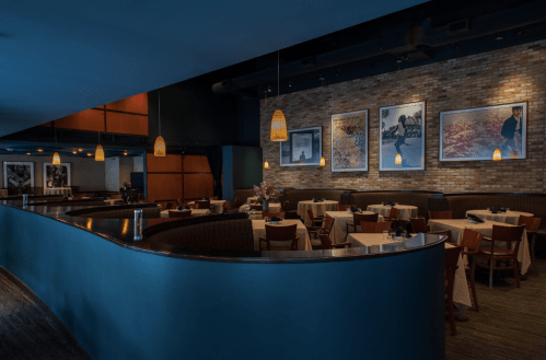 Interior of a restaurant featuring curved seating, tables set for dining, and framed artwork on the brick wall.