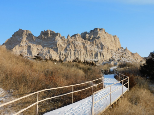 A snow-covered pathway leads through grass to rocky cliffs under a clear blue sky.