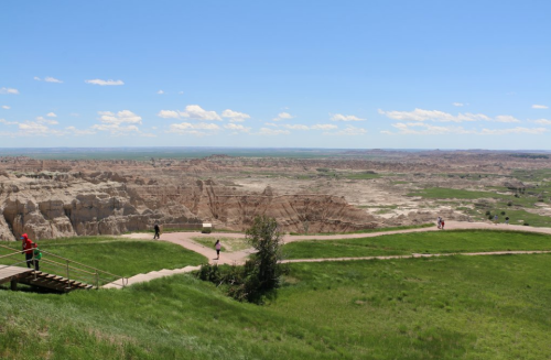 A panoramic view of a rugged landscape with green grass, cliffs, and people walking along a path under a blue sky.