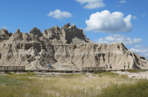 A scenic view of rugged rock formations under a blue sky with clouds, featuring people walking along a wooden path.