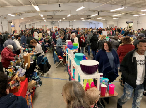 A crowded indoor market with numerous vendors and shoppers browsing various items. Colorful displays and merchandise are visible.