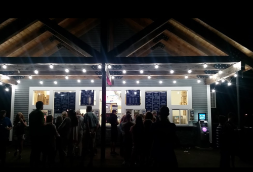 A crowd gathers outside a brightly lit food stand with a menu board under a wooden roof at night.