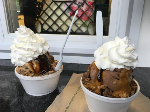 Two cups of ice cream topped with whipped cream and drizzled with chocolate and caramel sauces, placed on a table.