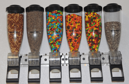 A row of six clear dispensers filled with various candies and toppings, mounted on a wall.