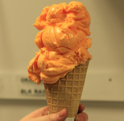 A hand holding a large cone of orange ice cream with a textured waffle cone.