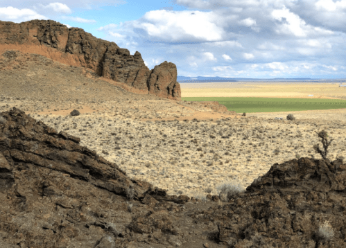 A rocky landscape with distant hills and a vast, open field under a partly cloudy sky.