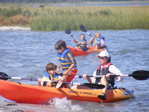 Two kayaks on a calm waterway, with children and adults paddling and enjoying a sunny day outdoors.