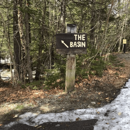 Wooden sign pointing left to "The Basin," surrounded by trees and a snowy path.