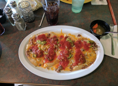 A plate of nachos topped with ground beef, cheese, salsa, and jalapeños, alongside a drink and a small bowl of pickled vegetables.