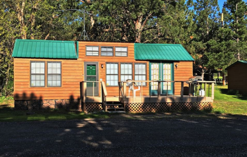A cozy wooden cabin with a green metal roof, featuring a front porch and surrounded by trees.