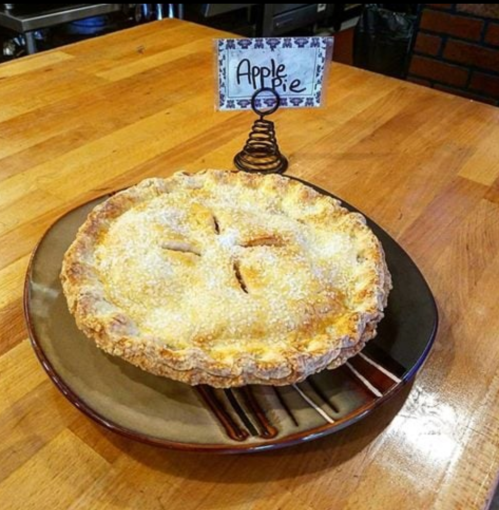 A freshly baked apple pie on a brown plate, displayed on a wooden table with a sign labeled "Apple Pie."