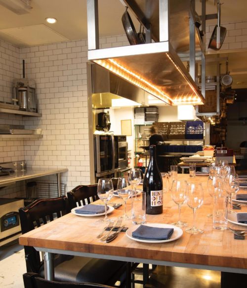 A modern kitchen with a wooden dining table set for a meal, featuring glassware and a wine bottle under warm lighting.