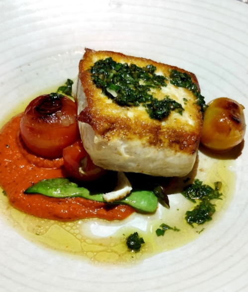 A piece of grilled fish topped with herbs, served on a bed of red sauce with vegetables and drizzled with olive oil.
