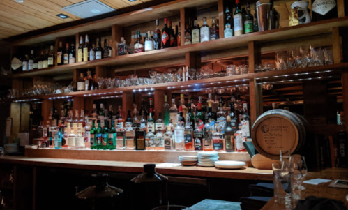 A well-stocked bar with various bottles of liquor, glasses, and a wooden barrel on a shelf.