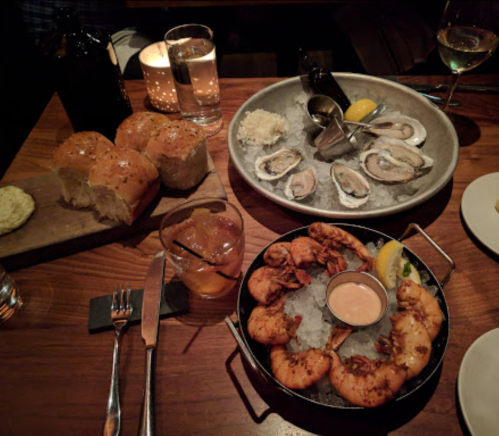 A table setting with shrimp on ice, oysters, bread rolls, and drinks in a dimly lit restaurant.