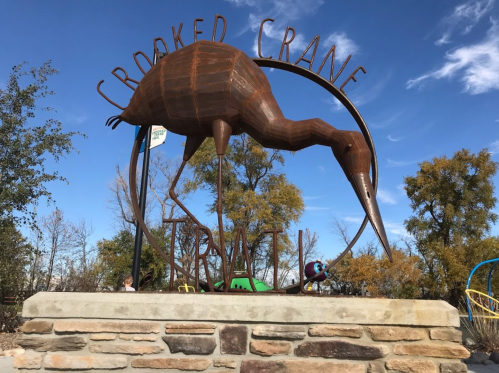A large metal sculpture of a crane stands atop a stone base, with "Crooked Crane" inscribed above it against a blue sky.