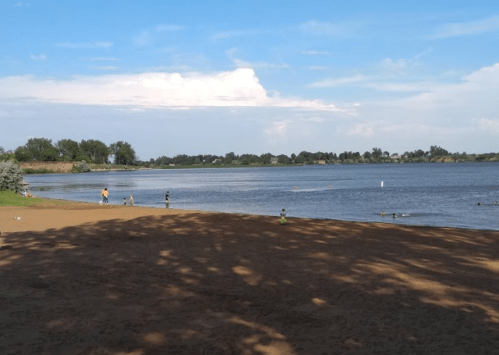 A sandy beach by a calm river, with people enjoying the water and trees lining the shore under a blue sky.