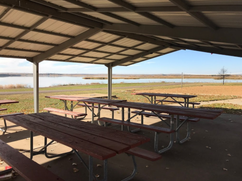 A covered picnic area with red tables overlooking a calm lake and grassy shoreline under a clear blue sky.