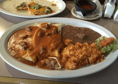A plate of enchiladas topped with red sauce, served with rice, refried beans, lettuce, and tomato.