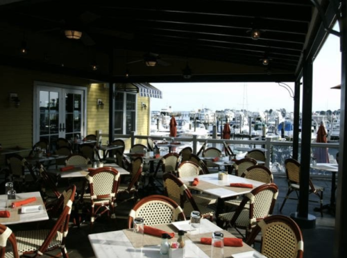Outdoor dining area with empty tables and chairs, overlooking a marina with boats in the background.
