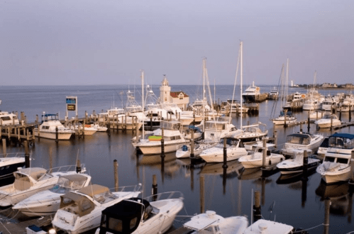 A serene marina at dusk, filled with various boats and a lighthouse in the background, reflecting on calm waters.